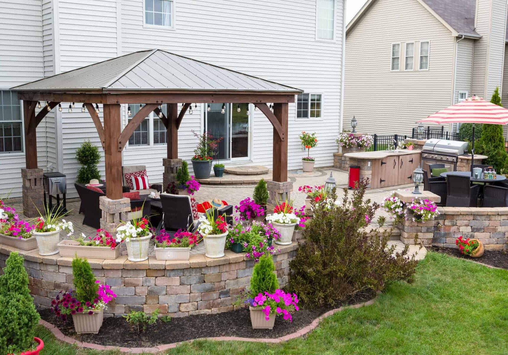 Colorful exterior curved patio with summer flowers and comfortable wicker armchairs under a covered wooden gazebo with aluminum roof in a neat upscale backyard